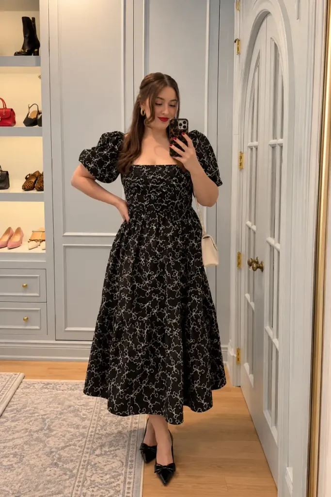 Woman wearing a black embroidered midi dress with puff sleeves, black bow heels, gold earrings, and a small ivory handbag for an elegant Christmas dinner outfit.