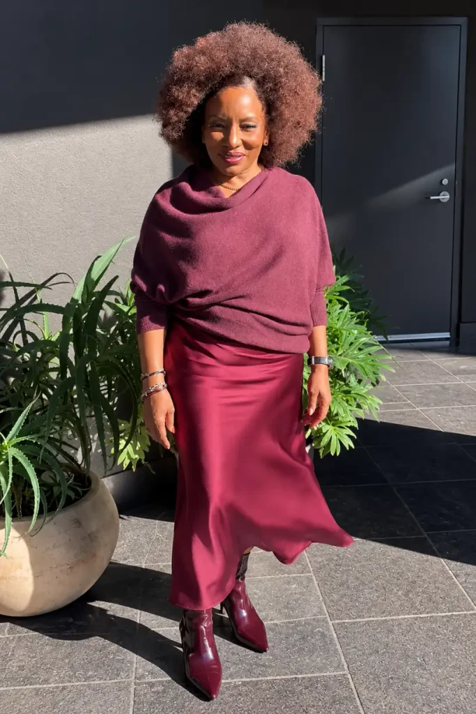 Black woman wearing a burgundy cowl-neck sweater with a matching satin midi skirt and glossy pointed ankle boots for an elegant fall outfit.