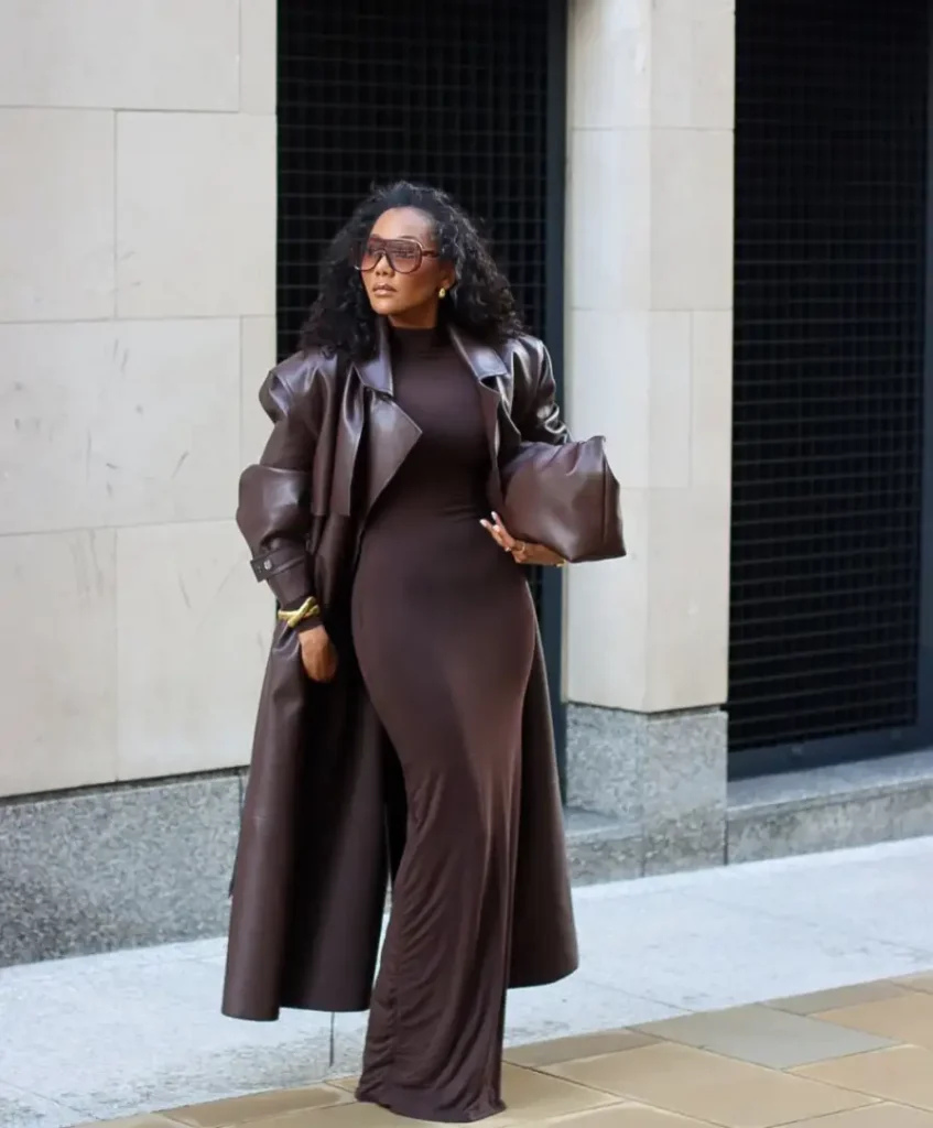 Black woman wearing a fitted chocolate brown maxi dress with a long brown leather trench coat, oversized sunglasses, gold jewelry, and a matching brown bag for a polished fall look.