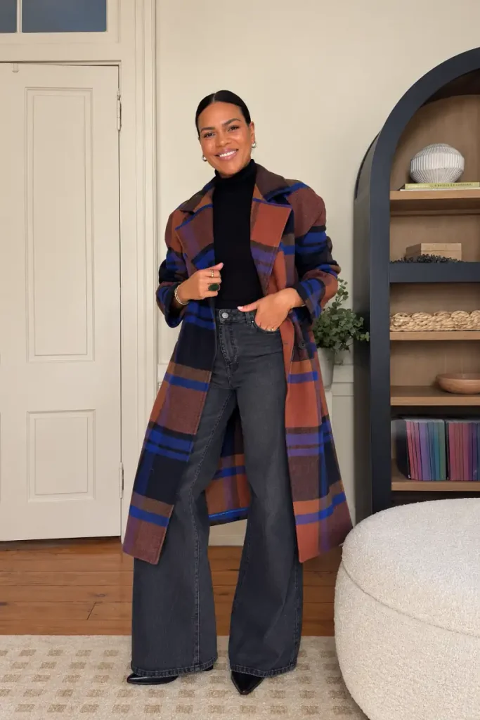 Black woman wearing a rust and navy plaid long coat over a black turtleneck, charcoal wide-leg jeans, and pointed black shoes for a classy fall outfit.