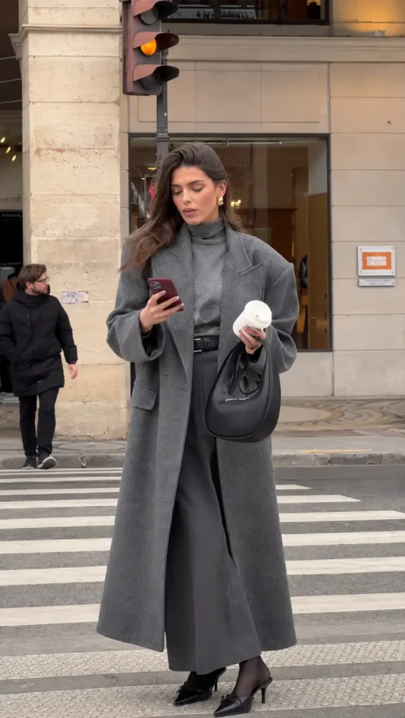 Woman wearing a long charcoal gray coat over a matching turtleneck and wide-leg trousers with black pointed heels and a black handbag for a polished fall city outfit.