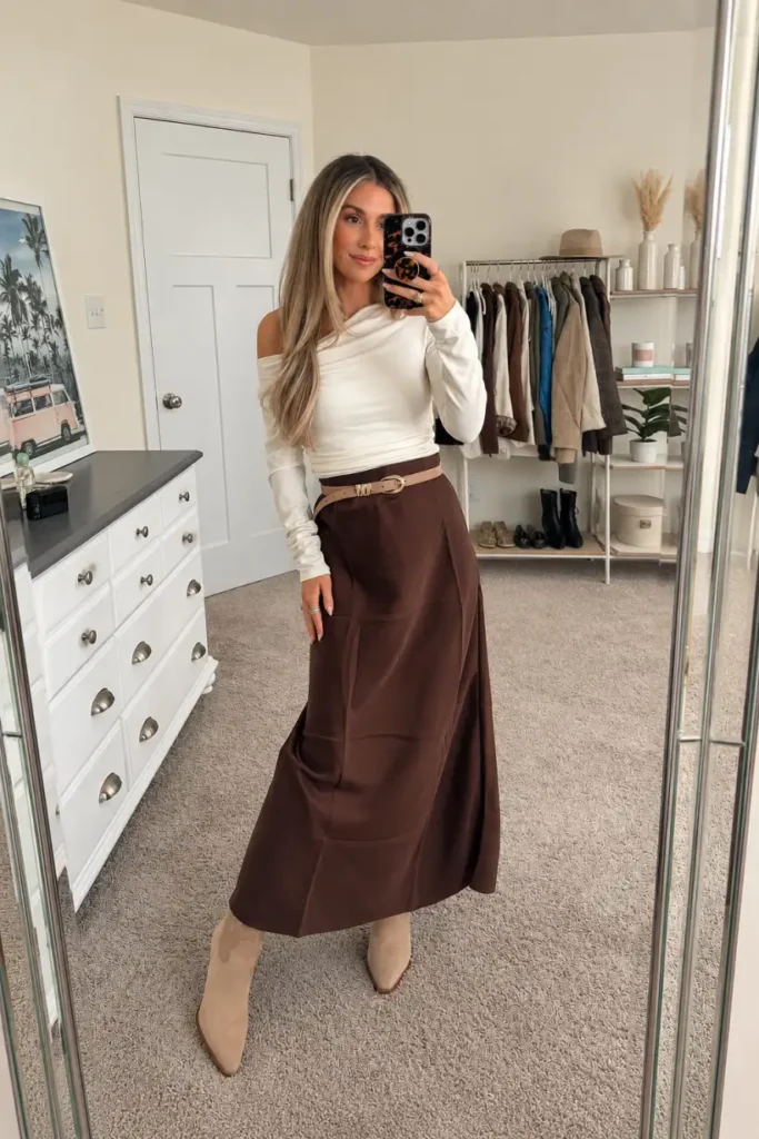Woman wearing an ivory off-shoulder top with a chocolate brown maxi skirt, tan waist belt, and beige cowboy boots for a classy Thanksgiving outfit.