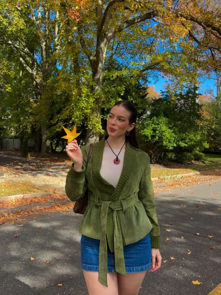 Fall outfit with olive belted jacket, cream top, denim mini skirt, and brown shoulder bag.