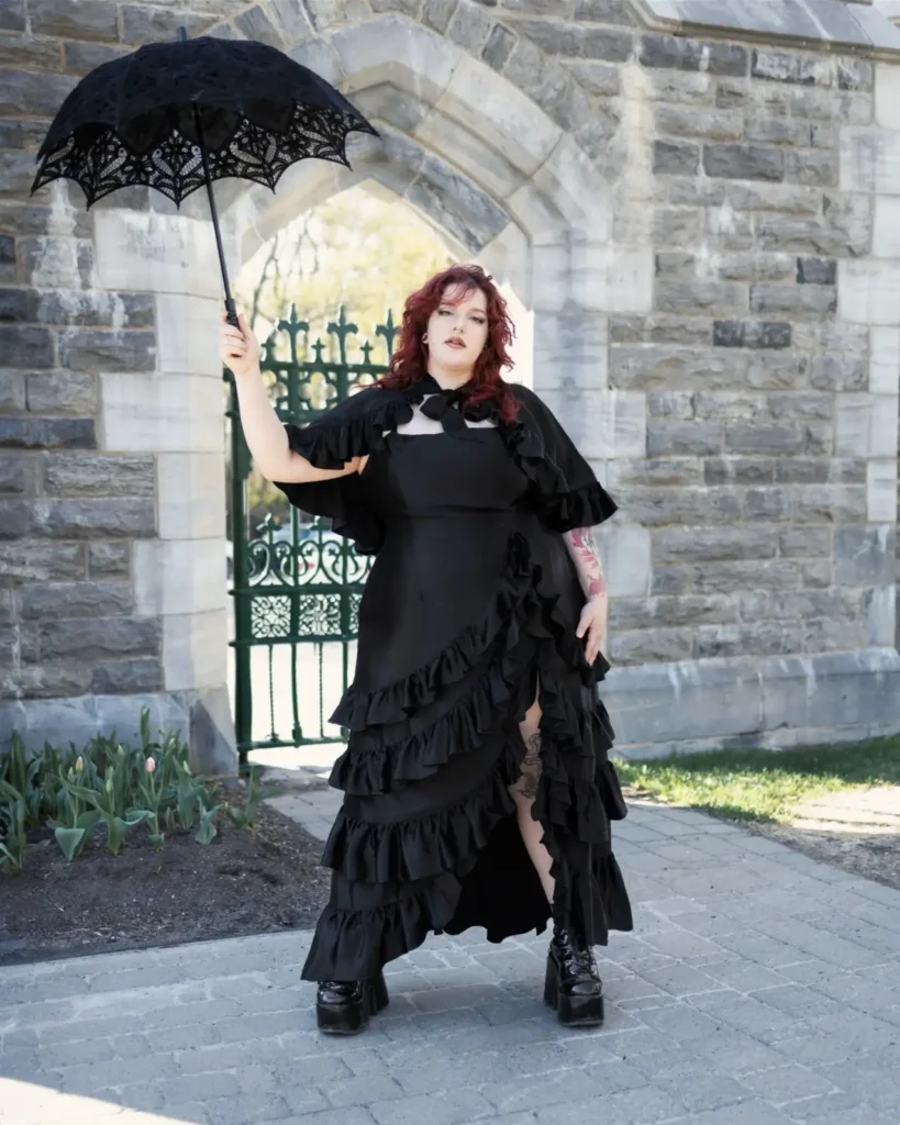 Plus size woman wearing a black Gothic Victorian witch dress with ruffles, platform boots, and a lace parasol.