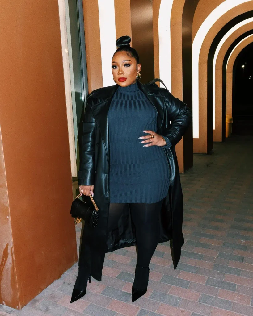 Plus size Black woman wearing a long black leather trench coat over a navy ribbed turtleneck, black leggings, pointed heels, and a small black handbag for a classy fall outfit.