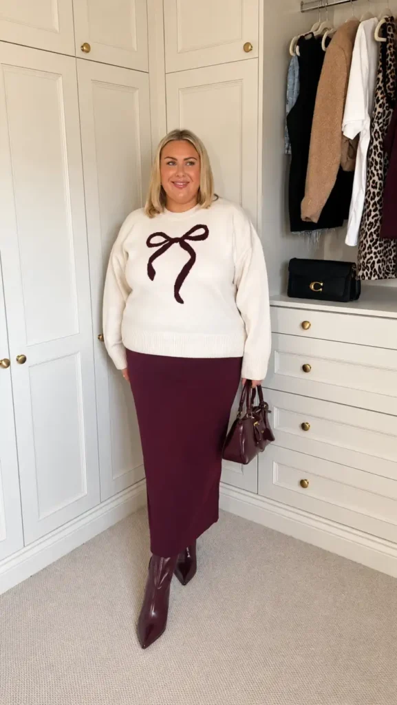 Plus size woman wearing a cream sweater with a burgundy bow, matching burgundy maxi skirt, pointed boots, and handbag for a feminine winter outfit.