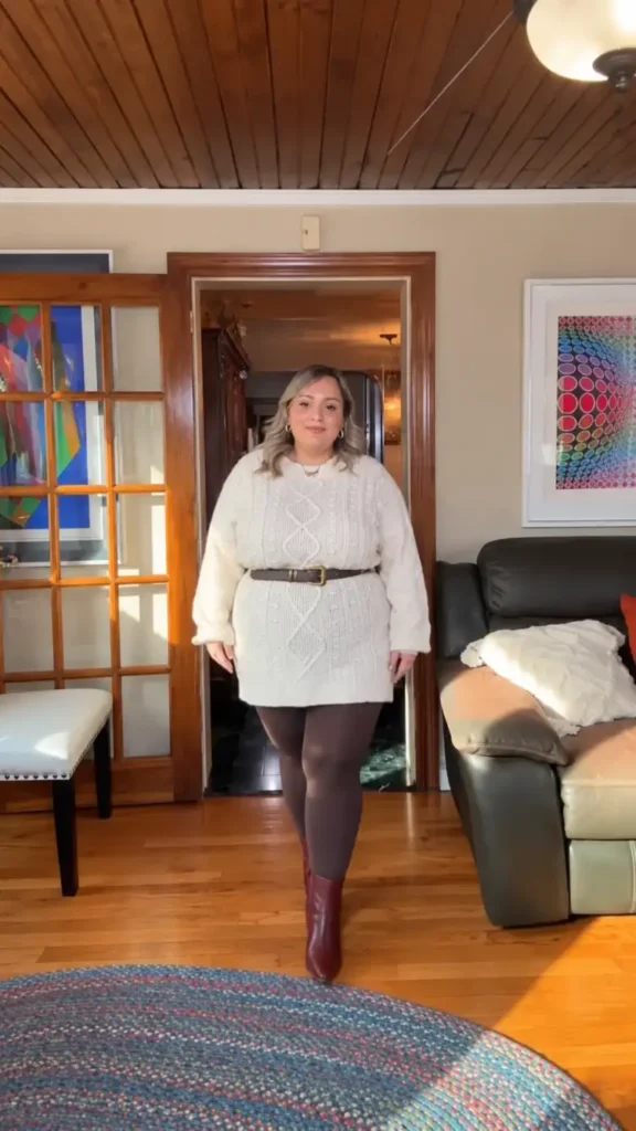 Plus size woman wearing a cream cable-knit sweater dress with a brown waist belt, chocolate tights, and burgundy boots for a cozy Thanksgiving outfit.