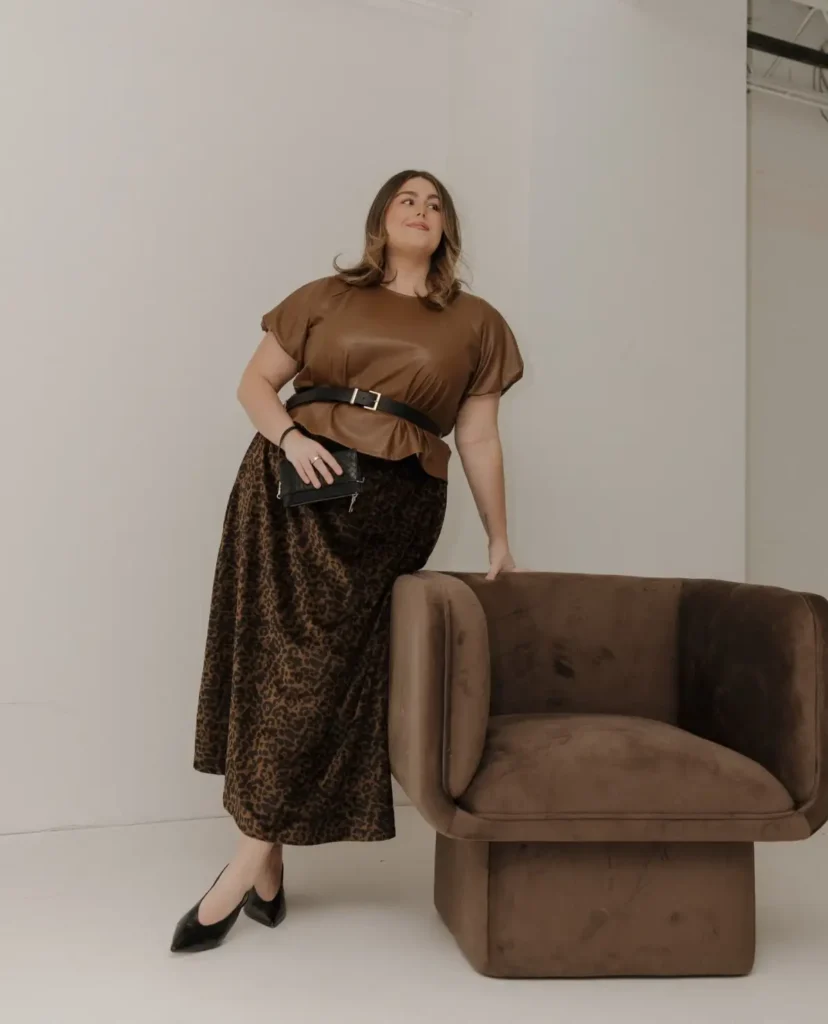 Plus size woman wearing a brown belted top with a leopard print midi skirt, black pointed flats, and a small black handbag for a polished fall outfit.
