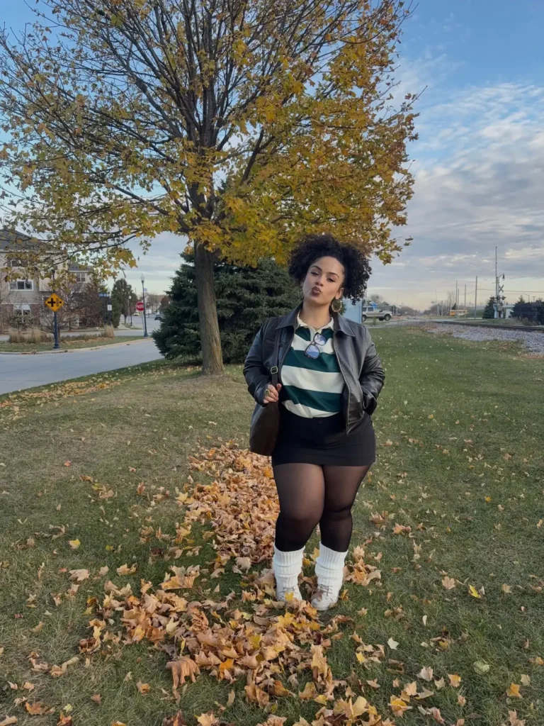 Plus size fall outfits Black woman wearing a striped rugby shirt, black leather jacket, mini skirt, sheer tights, white leg warmers, and sneakers for a casual fall outfit.