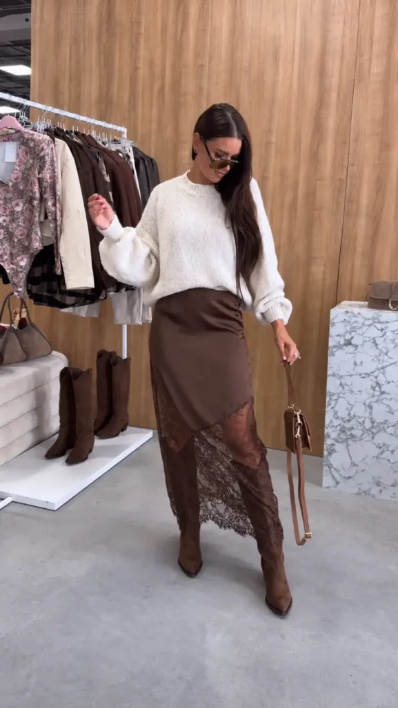 Woman wearing a cream oversized sweater with a brown lace midi skirt, matching brown boots, and a small handbag.