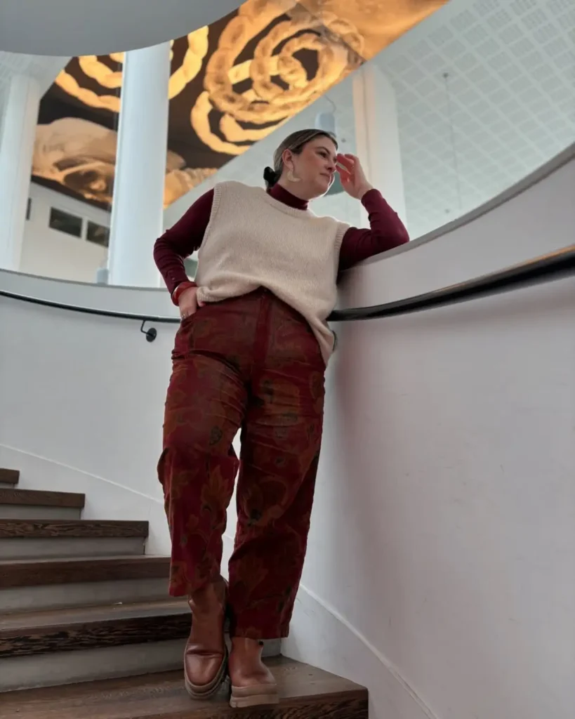 Plus size fall outfit with a cream sweater vest layered over a burgundy turtleneck, patterned burgundy pants, and brown ankle boots.