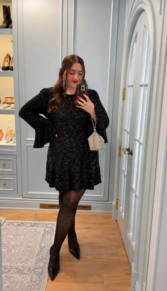 Woman wearing a black long-sleeve sequin mini dress with flared sleeves, sheer polka-dot tights, black pointed heels, and a crystal handbag for a holiday party.