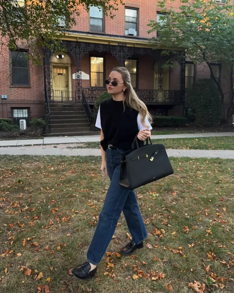 Woman wearing a black sweater vest over a white T-shirt with straight blue jeans, black loafers, and a structured black tote for a casual fall look.