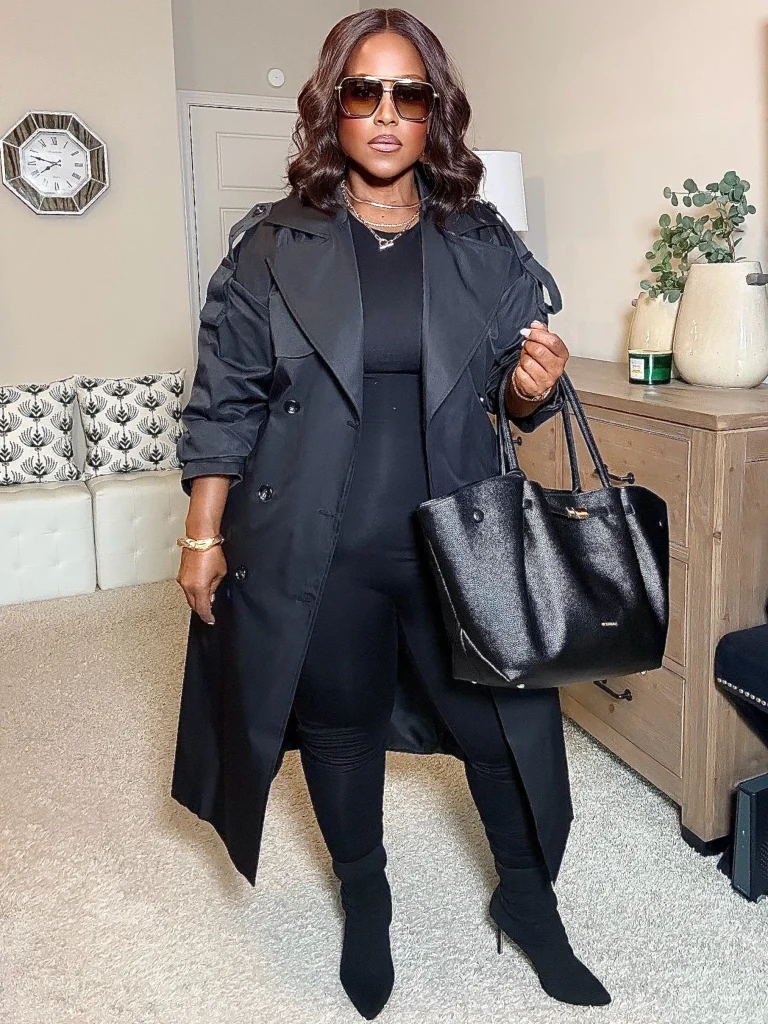 Black woman wearing a long black trench coat with a fitted black top, slim trousers, pointed ankle boots, and a large black tote for a sleek monochrome fall outfit.
