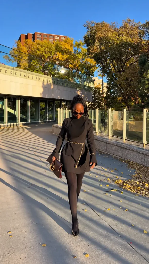 Black woman wearing a tailored black blazer mini dress with a gold chain belt, sheer black tights, pointed heels, and a burgundy clutch.