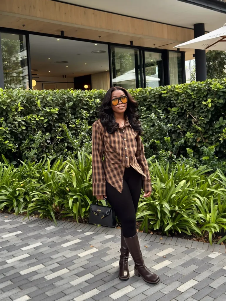 Black woman wearing a brown plaid long shirt with black fitted pants, tall brown riding boots, oversized sunglasses, and a structured black handbag for a casual fall look.