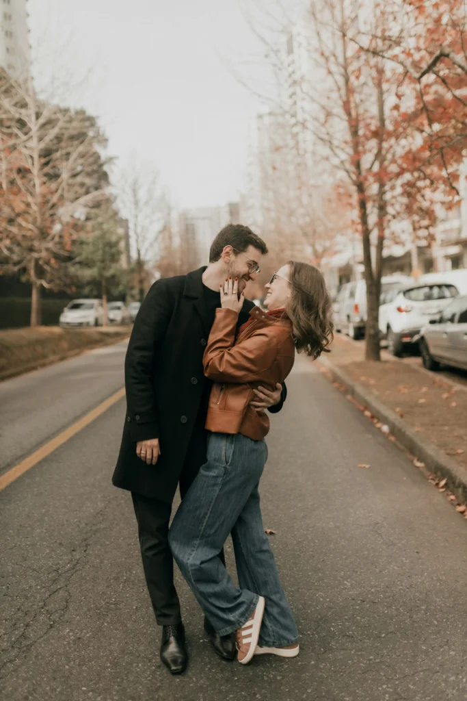 Couple wearing coordinating fall streetwear with a cognac leather-look jacket, wide-leg blue jeans, tan sneakers, long black coat, and dark tailored trousers.