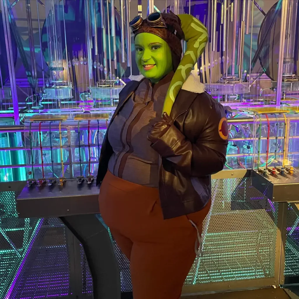 Plus size woman dressed as a green galactic pilot with fantasy face makeup, goggles, brown jacket, and sci-fi accessories.