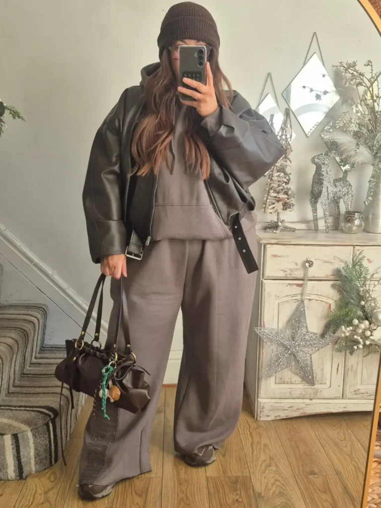 Plus size woman wearing a black oversized leather jacket over a taupe-gray hoodie and matching wide-leg sweatpants with a brown beanie and chunky sneakers.