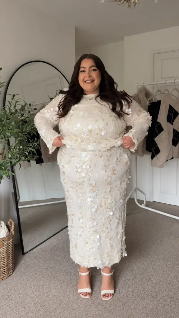 Plus-size woman wearing a matching ivory sequin long-sleeve top and fitted maxi skirt with oversized circular embellishments and white strappy heels for a glamorous holiday look.