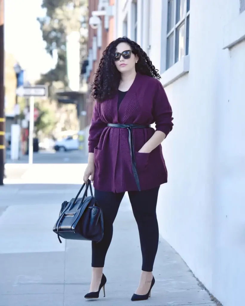 Plus size fall outfit with a long burgundy cardigan cinched with a slim black belt, black leggings, black pointed pumps, and a structured black handbag.