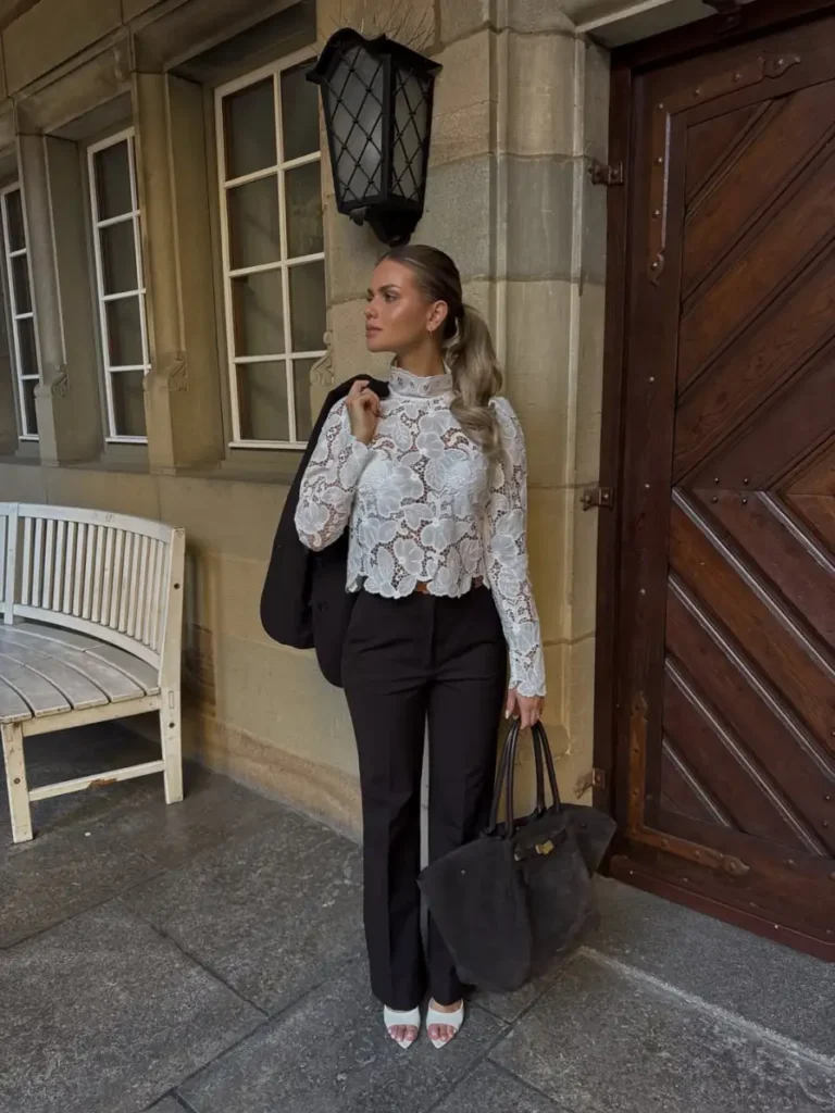 Woman wearing a high-neck white lace blouse with black tailored trousers, white heels, and a large dark handbag.