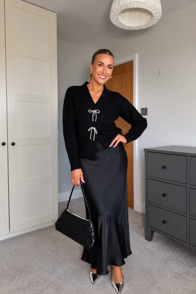 Woman wearing a black ribbed cardigan with silver bow details, a black satin maxi skirt, metallic pointed heels, and a woven handbag for an elegant holiday outfit.