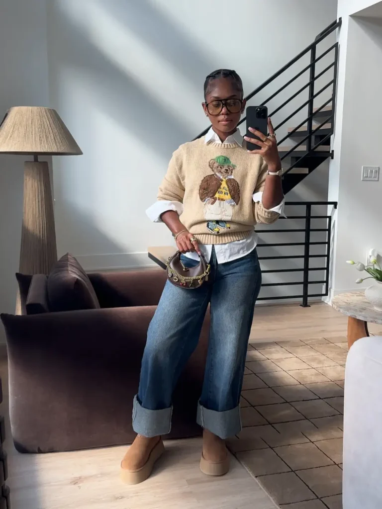 Black woman wearing a cream teddy bear sweater layered over a white button-down shirt with wide cuffed jeans, tan platform shoes, and a small brown handbag for a preppy fall outfit.
