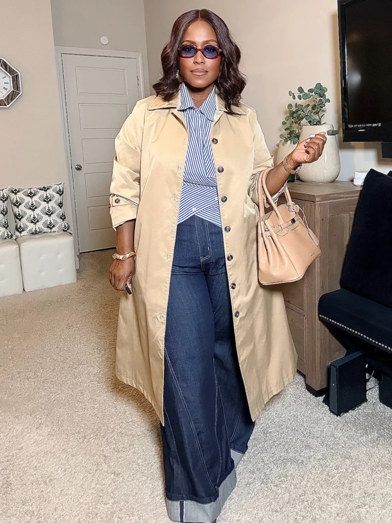Black woman wearing a beige trench coat over a blue-and-white striped shirt with dark wide-leg cuffed jeans and a blush handbag for a casual autumn look.