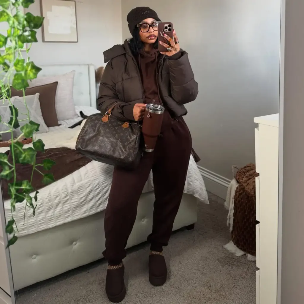 Black woman wearing a chocolate brown hoodie and matching joggers with a cropped brown puffer jacket, knit beanie, platform clogs, and patterned handbag.