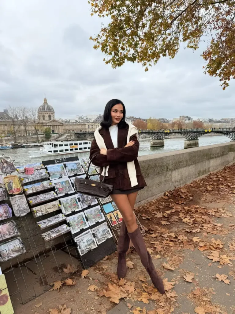Woman wearing a chocolate brown jacket with matching shorts, a white top, cream sweater over the shoulders, and tall brown boots for a Paris-inspired fall outfit.