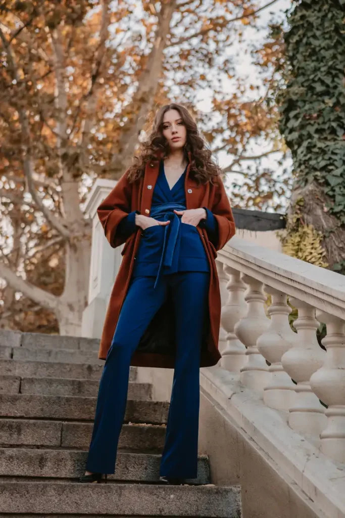 Woman wearing a cobalt blue wrap-style top with matching tailored trousers, a rust brown long coat, and black heels for a classy Thanksgiving dinner look.