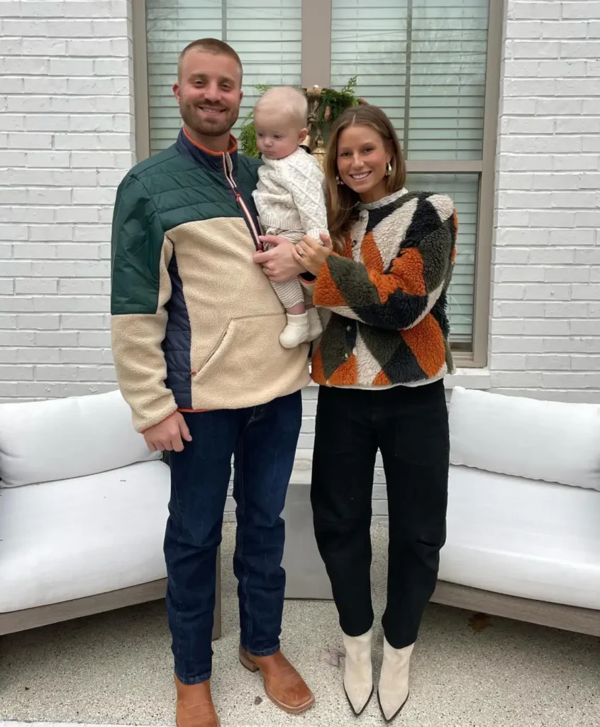Family wearing cozy fleece outfits with Dad in a green and cream pullover, Mom in an orange and olive colorblock sherpa jacket, and baby in a cream knit sweater and striped pants.