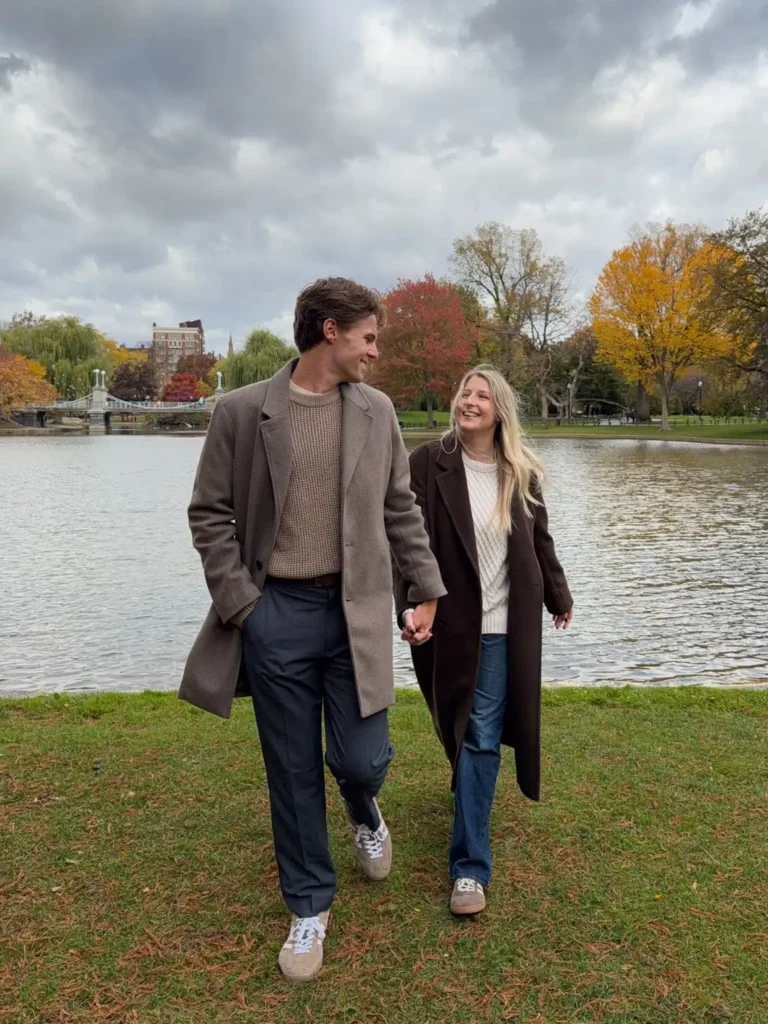 Couple walking beside a lake wearing taupe and dark brown long coats, beige and cream knit sweaters, navy trousers, blue jeans, and neutral sneakers.