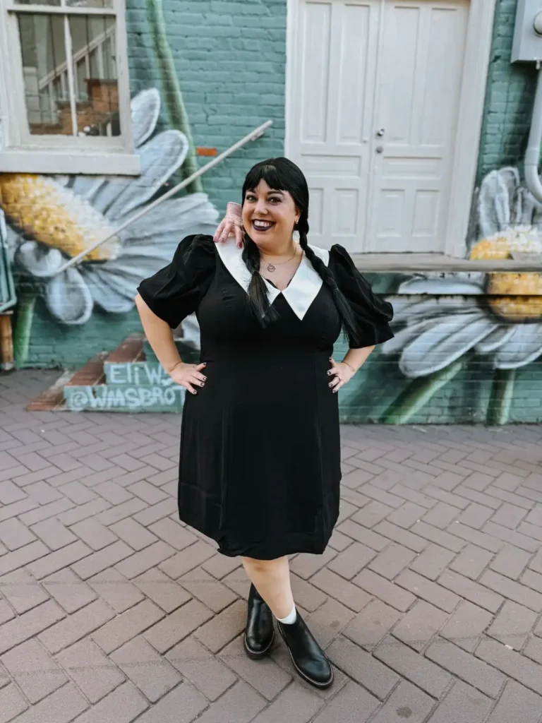 Plus size woman wearing a black Gothic schoolgirl dress with a white collar, braids, ankle socks, and black boots.