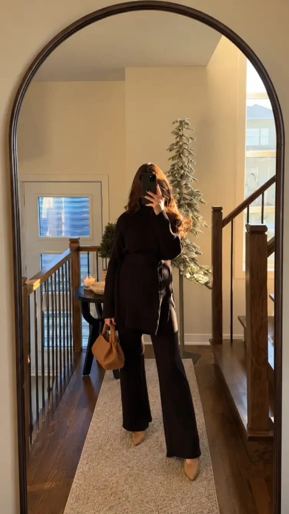 Pregnant woman wearing a black ribbed belted tunic sweater with matching wide-leg knit pants, neutral pointed heels, and a tan handbag for a classy maternity outfit.