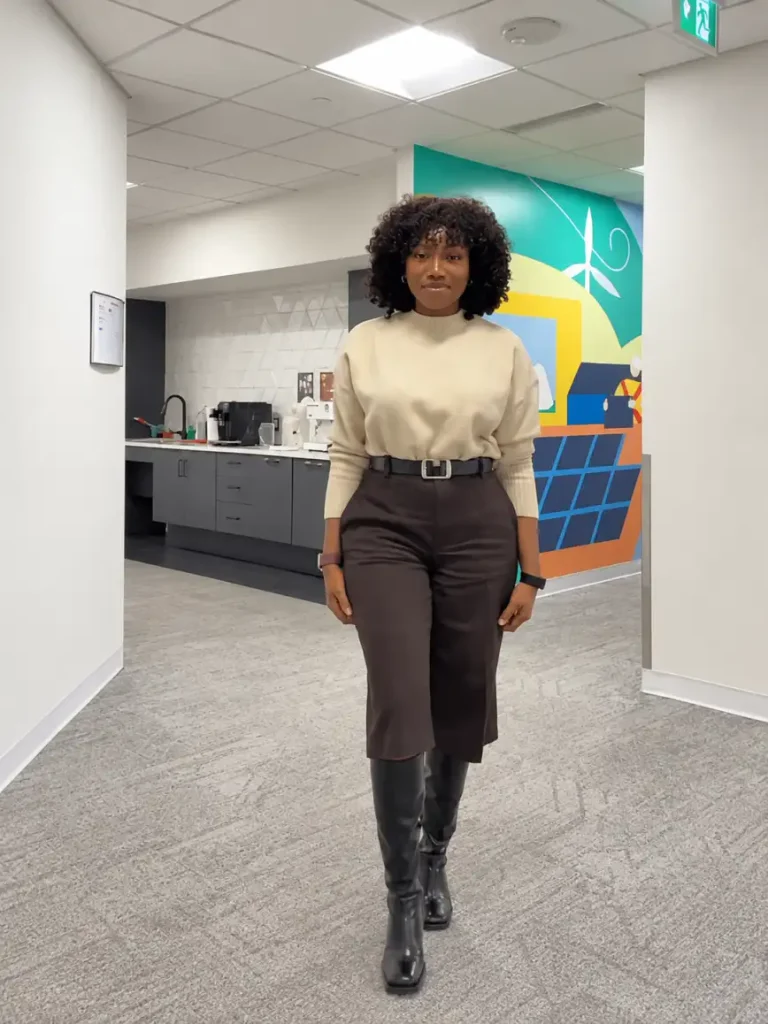 Black woman wearing an oatmeal high-neck sweater with a dark brown belted midi skirt and tall black boots for a cozy, professional autumn office look.