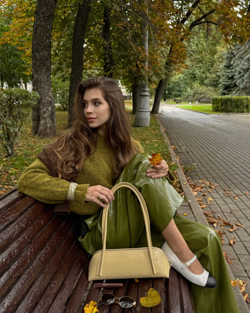 Woman wearing an olive green sweater with matching leather-look trousers, cream Mary Jane flats, and a light beige handbag for a tonal fall outfit.