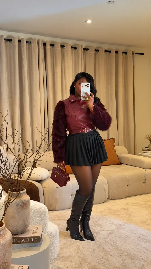Plus size Black woman wearing a burgundy leather-look top with faux-fur sleeves, black pleated mini skirt, sheer tights, and knee-high boots for a stylish fall outfit.