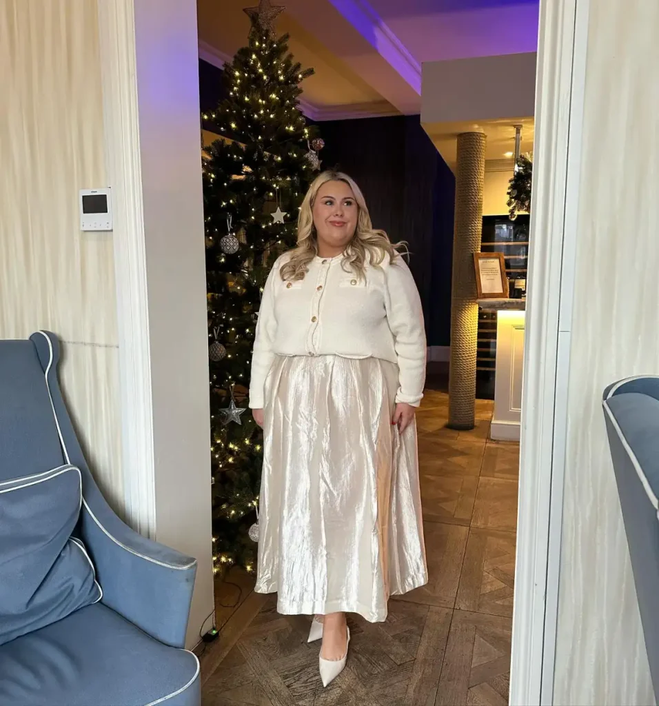 Plus-size woman styling a cream gold-button cardigan with a flowing champagne satin maxi skirt and ivory pointed heels for an elegant Christmas dinner outfit.