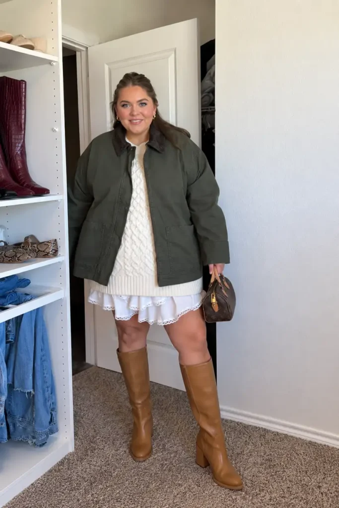 Plus size woman wearing an olive green barn jacket over a cream cable-knit sweater and white ruffled skirt, styled with tan knee-high boots.