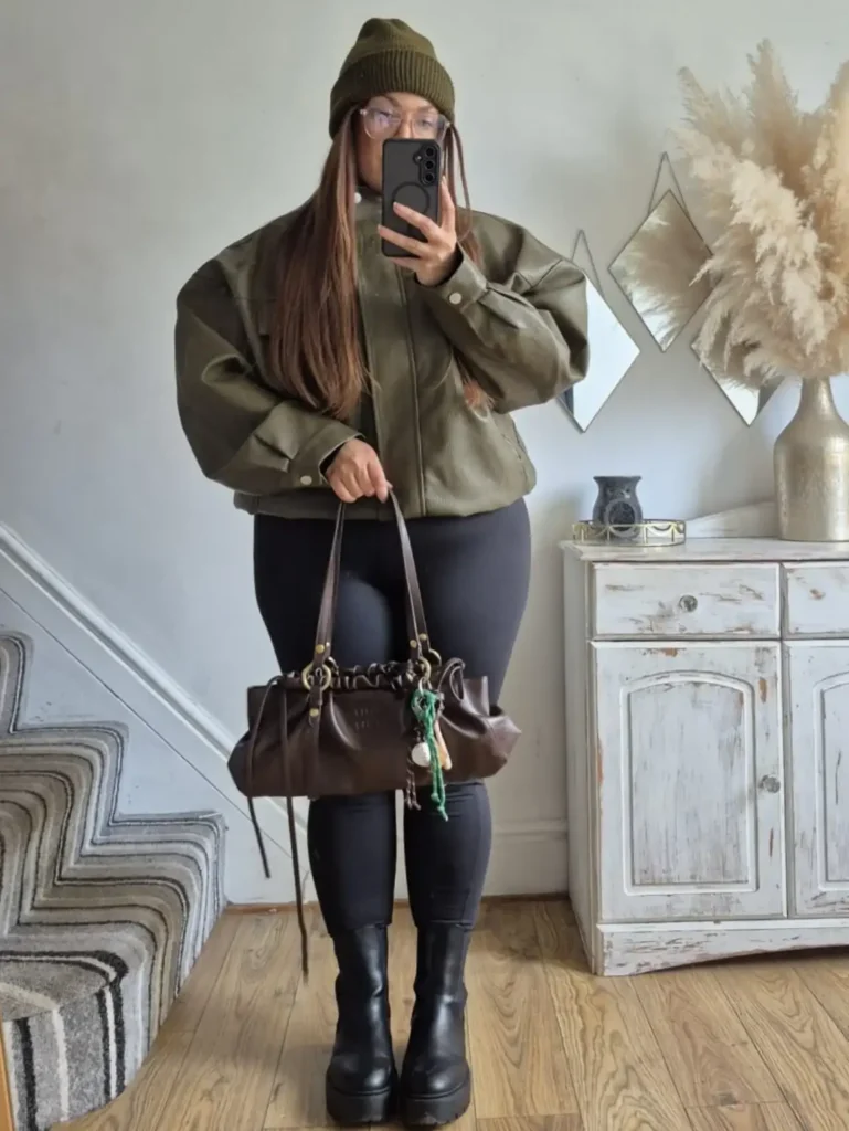 Plus size woman wearing an olive green leather bomber jacket with black leggings, chunky knee-high boots, an olive knit beanie, and a dark brown handbag.