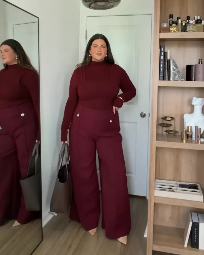 Plus size woman wearing a burgundy turtleneck with matching high-waisted wide-leg trousers, pointed heels, and a dark tote bag for polished fall workwear.