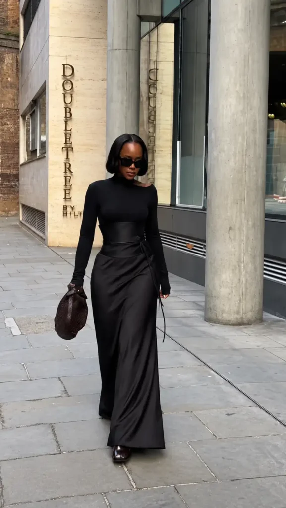 Black woman wearing a fitted black long-sleeve top with a sculpted waist detail and flowing black maxi skirt, styled with sunglasses and a woven brown clutch.