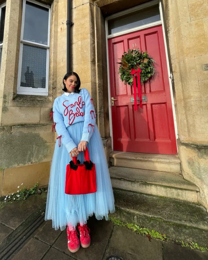 Woman wearing a light blue festive sweater with red sleeve bows, a matching layered tulle maxi skirt, bright red sneakers, and a red handbag for a playful Christmas outfit.