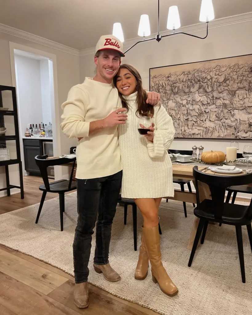 Couple wearing cozy Thanksgiving dinner outfits with an ivory cable-knit sweater dress, caramel knee-high boots, cream sweatshirt, dark jeans, and tan suede boots.