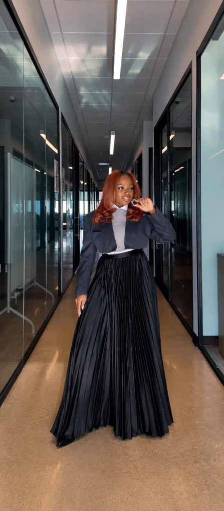 Black woman styling a cropped charcoal blazer over a light top with a long black pleated maxi skirt and pointed shoes for an elegant creative office outfit.