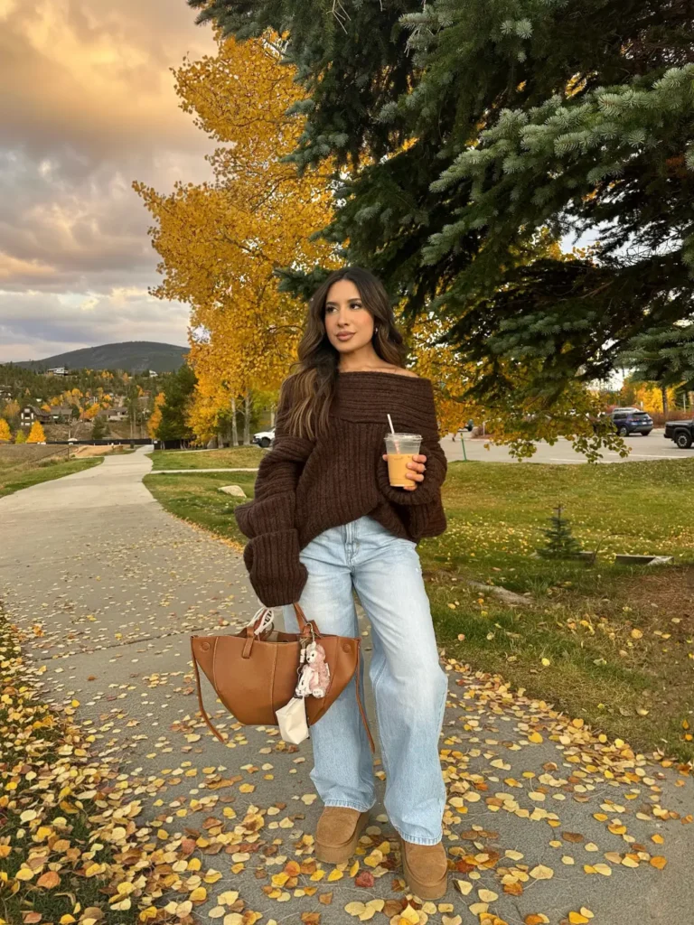 Woman wearing a chunky espresso brown off-shoulder sweater with light-wash baggy jeans, chestnut platform slip-on shoes, and a tan tote bag for a cozy autumn outfit.