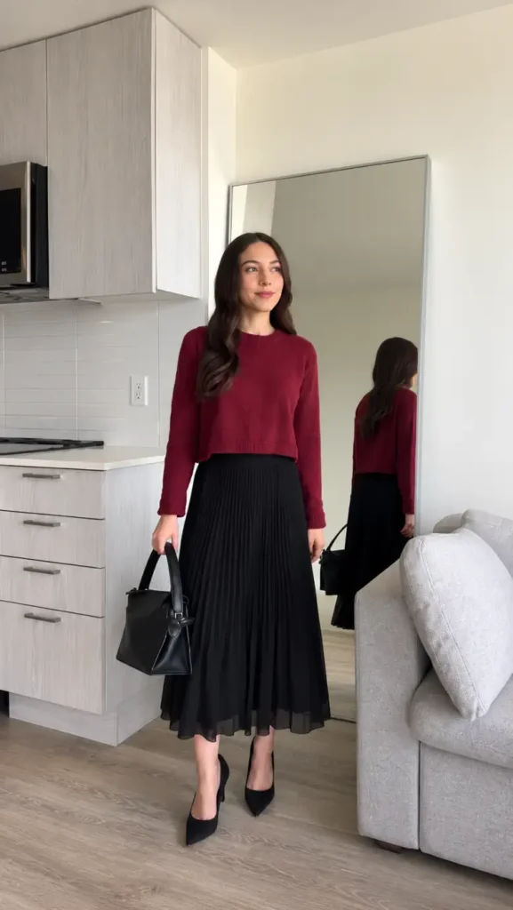 Petite woman wearing a cropped burgundy sweater with a black pleated midi skirt, pointed black pumps, and a structured handbag for fall.