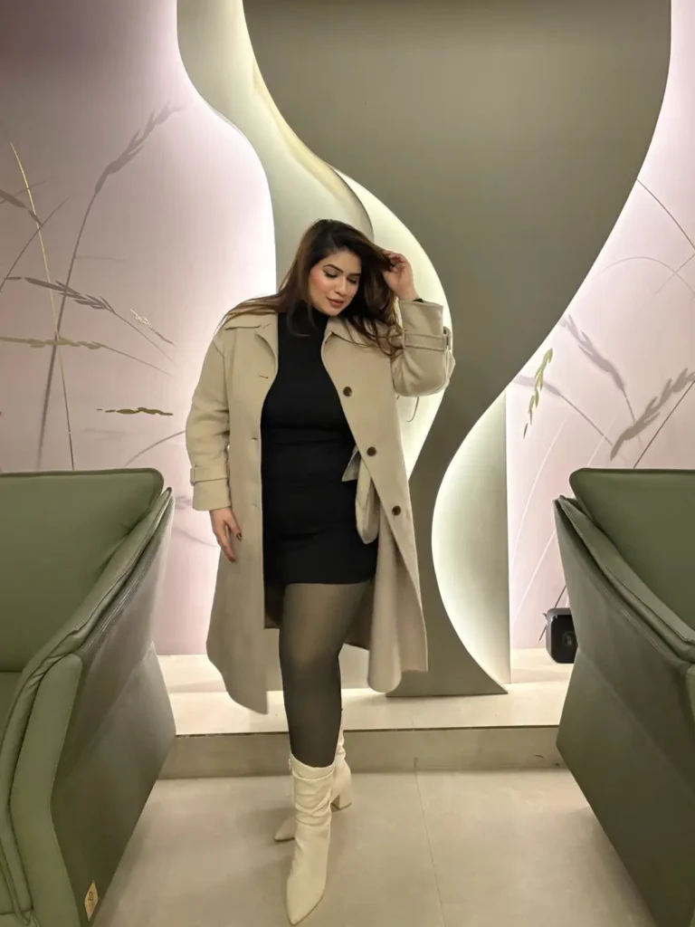 Plus size woman styling a long beige winter coat over a fitted black mini dress with sheer tights and cream knee-high heeled boots for a classy winter look.
